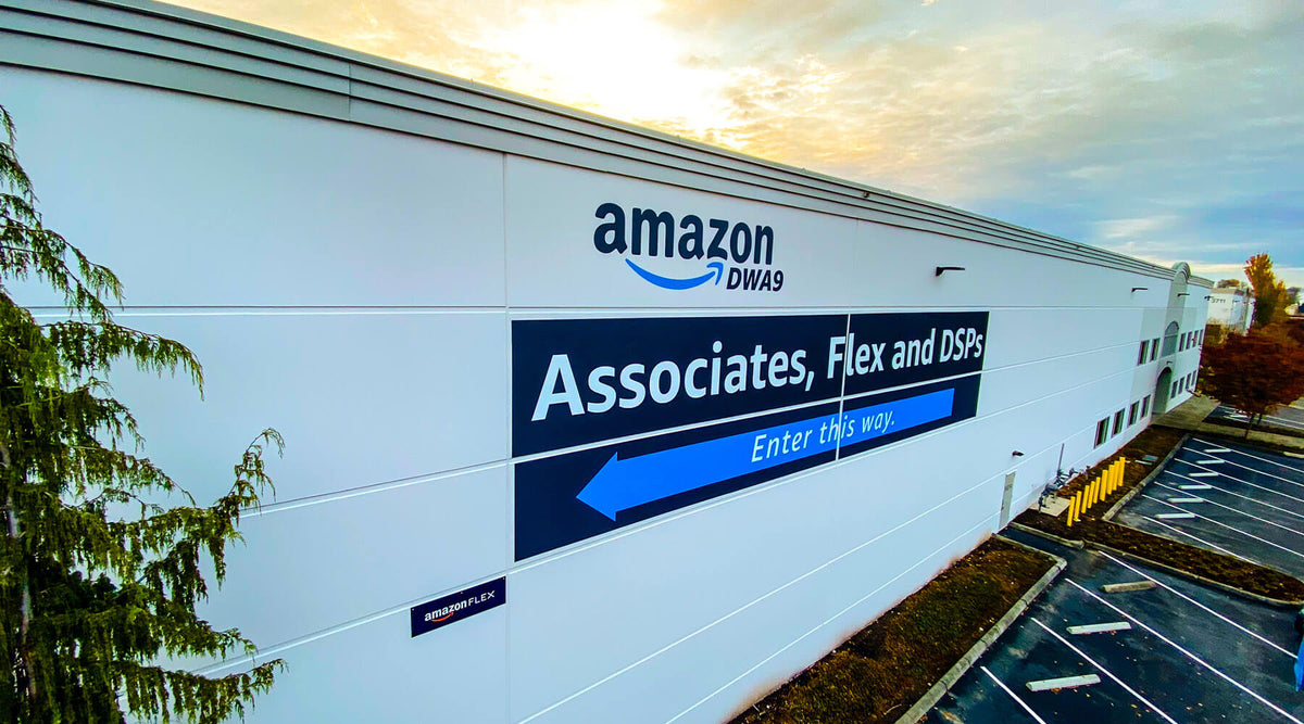 Dark blue Amazon building graphic with a directional arrow on a white warehouse wall at sunset