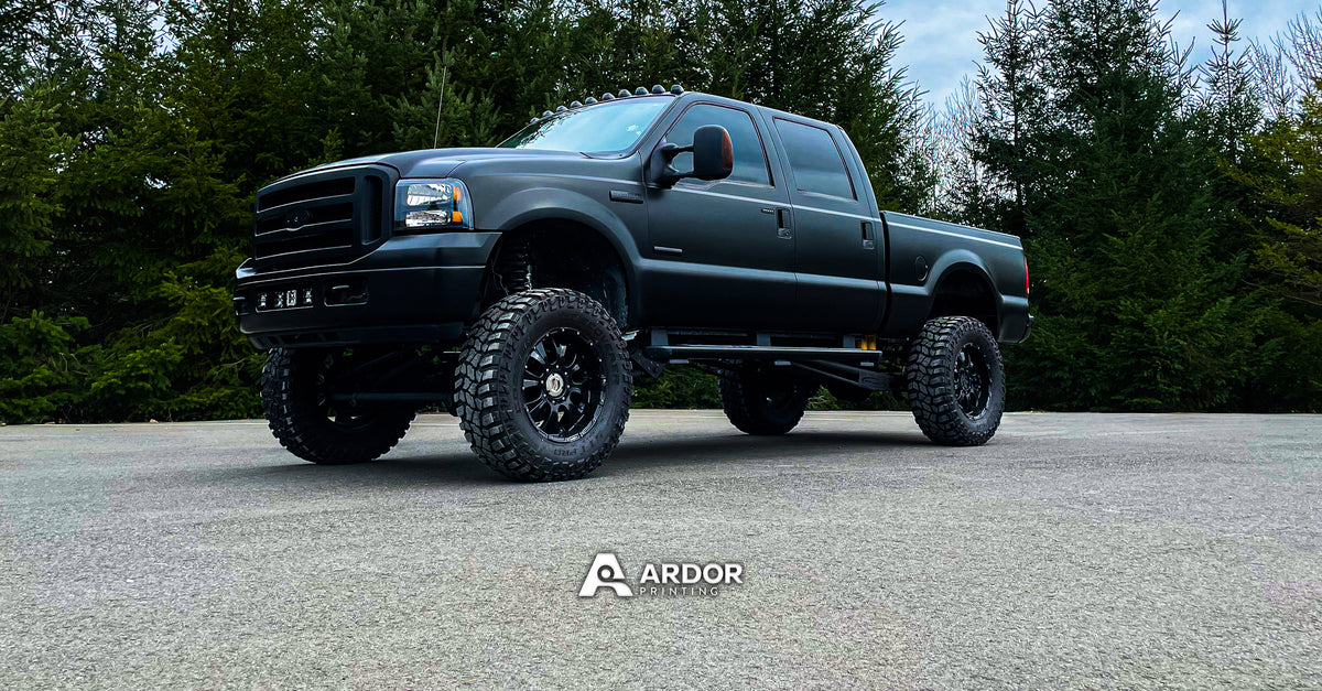 Lifted Ford F-250 pickup wrapped in satin black vinyl, parked in front of evergreen trees