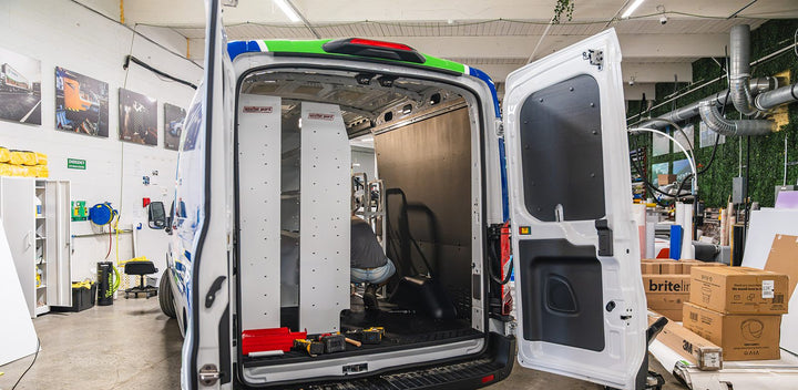 White work van with rear doors open in the Ardor Printing shop bay, surrounded by tools, boxes and printed photo displays
