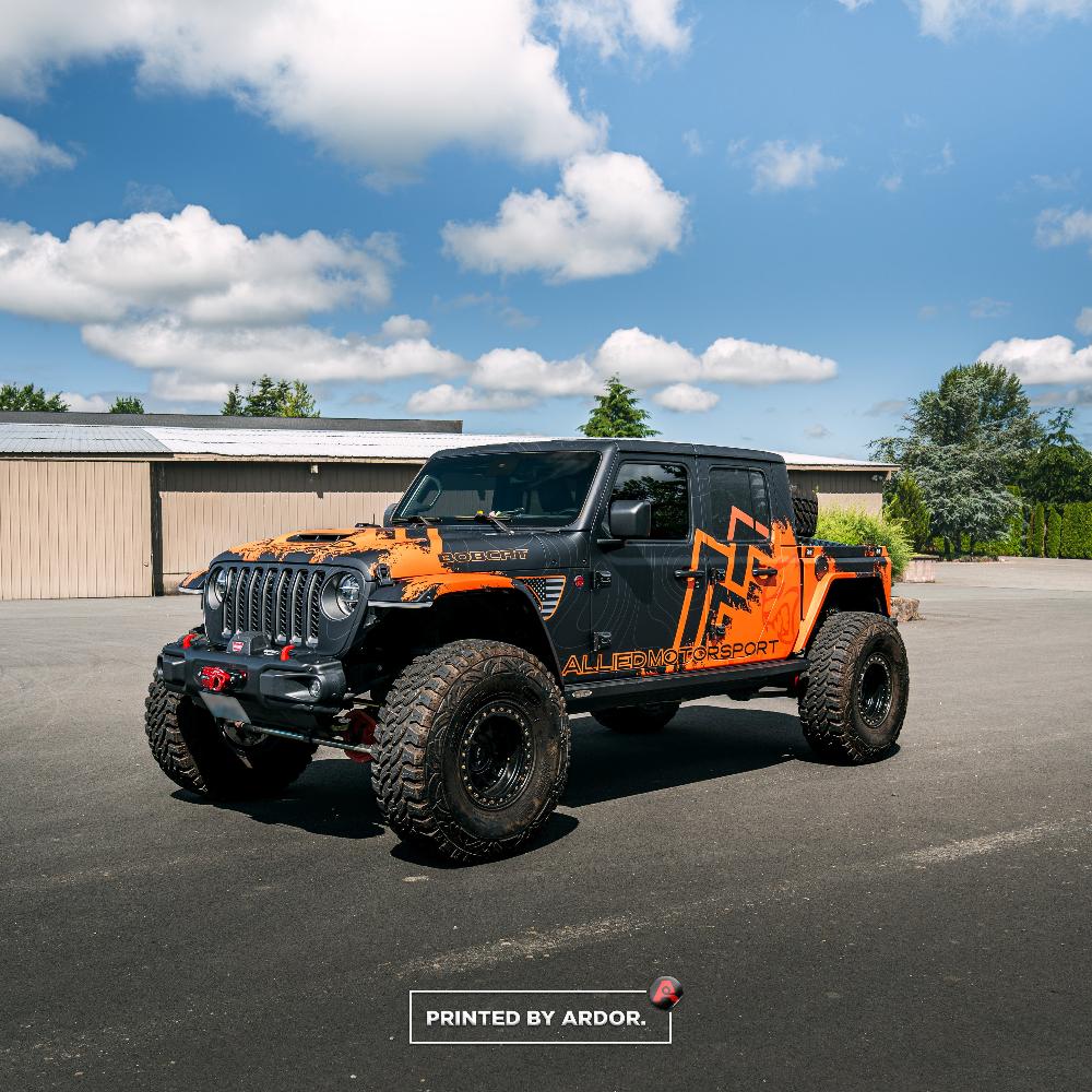 Allied Motorsport Jeep truck with a custom black and orange wrap, featuring off-road tires and branding, parked on asphalt under a clear sky.