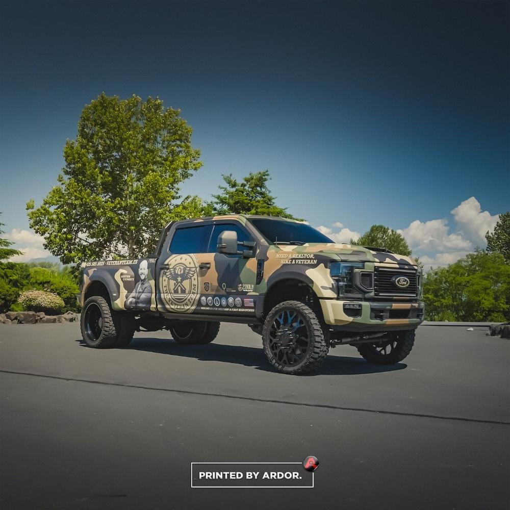 Ford F450 truck with a camouflage wrap, featuring veteran and realtor branding, parked outdoors under a clear sky with trees in the background.