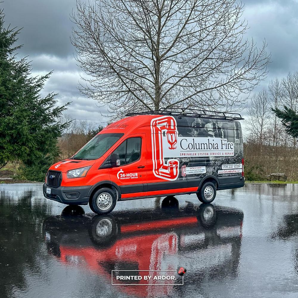 Columbia Fire van with a red and black wrap, featuring fire sprinkler system services, emergency contact info, and company branding, parked on wet pavement.