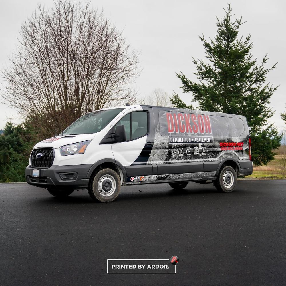Dickson Demolition van with a black, white, and gray custom wrap, featuring company services and contact info, parked near trees on a paved road.