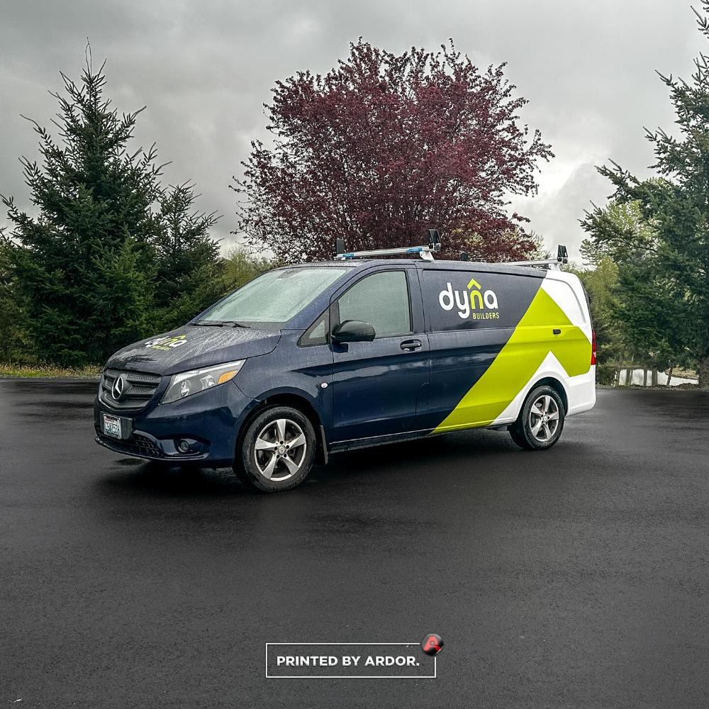 Dyna Builders van with a blue, white, and yellow custom wrap featuring company branding, parked on a wet road with trees in the background.