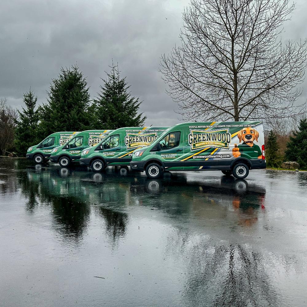 A lineup of four Greenwood Heating vans wrapped in green and yellow, featuring company branding and a dog mascot, parked on a rainy day.