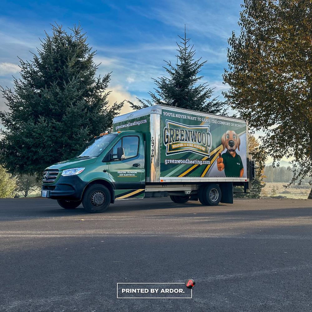 Custom-wrapped Greenwood Heating truck in green and white, displaying company logo, website, and mascot, parked on a road surrounded by trees.