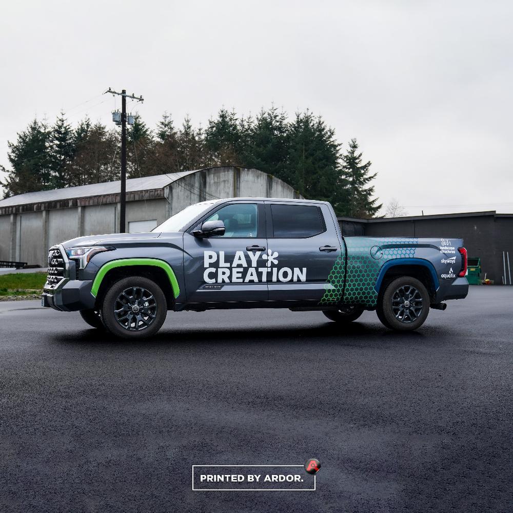 Play Creation truck with a gray and green custom wrap, featuring company branding and logo, parked on a paved road near an industrial building.