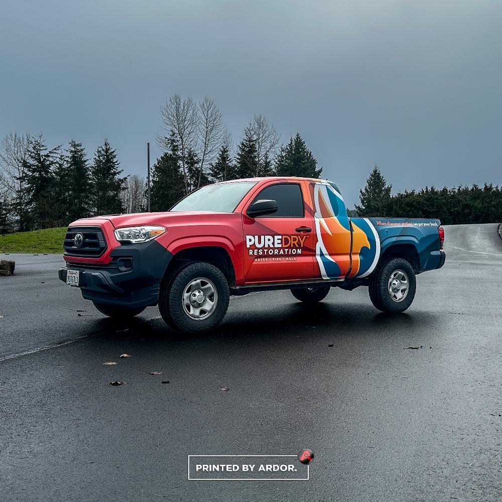 PureDry Restoration truck with a red, orange, and blue custom wrap, showcasing company branding and parked on a wet road near trees.