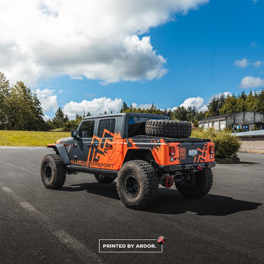 Rear-side view of an Allied Motorsport Jeep Gladiator with a black and orange custom wrap, oversized tires, and a spare tire mounted in the truck bed.
