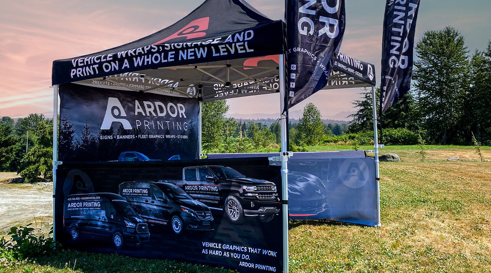A vibrant and eye-catching custom event tent setup by Ardor Printing, featuring a full-color printed canopy, back wall, side half walls, and two feather flags, all in dark colors with black, gray, and maroon accents, on a grass lawn during a sunny day.