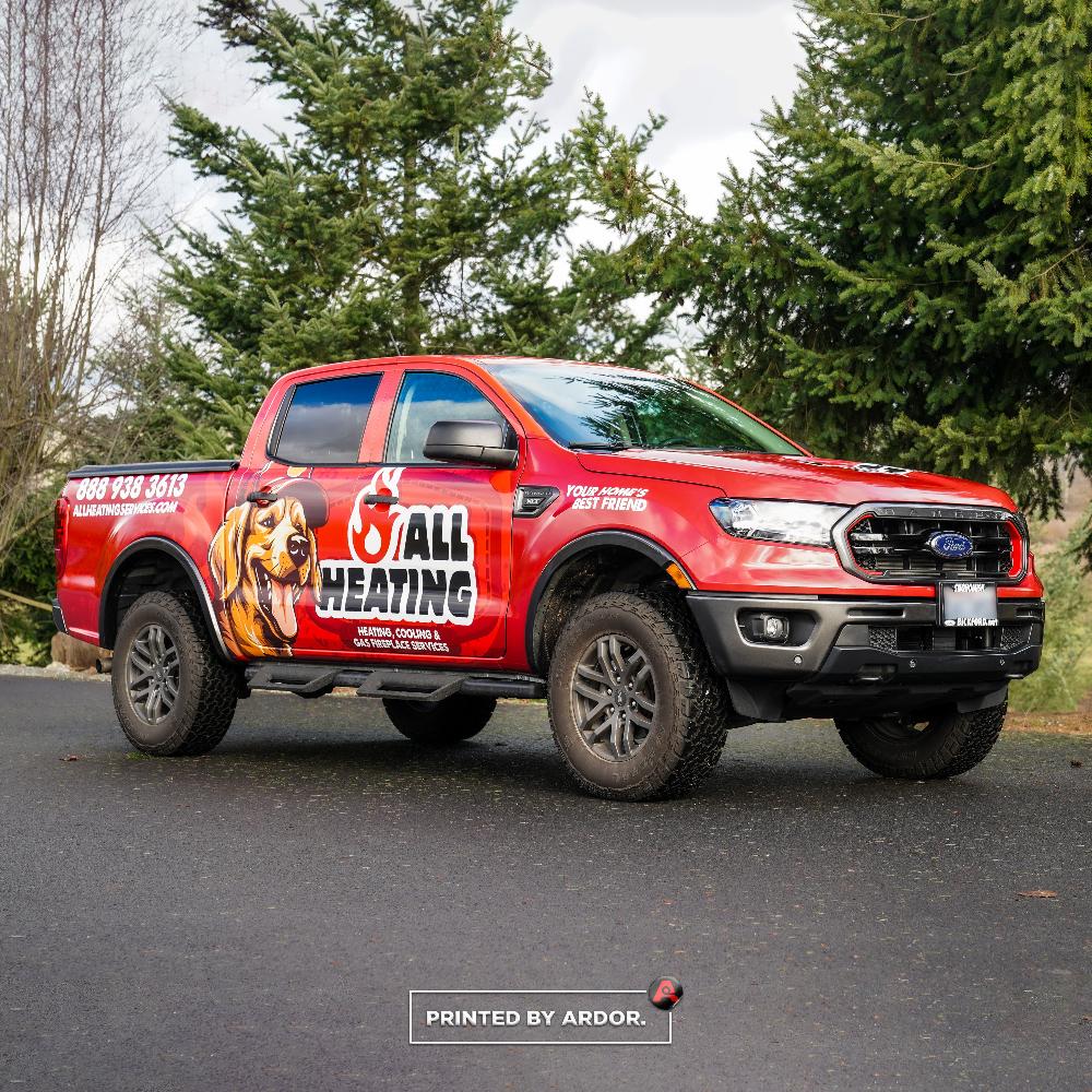 Custom-wrapped All Heating truck in red, showcasing company logo, dog mascot, and contact info, parked on a paved road surrounded by trees.