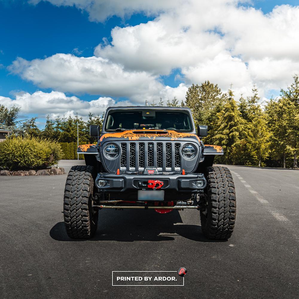 Front view of an Allied Motorsport Jeep with a black and orange custom wrap, featuring large off-road tires and a rugged bumper, parked outdoors.