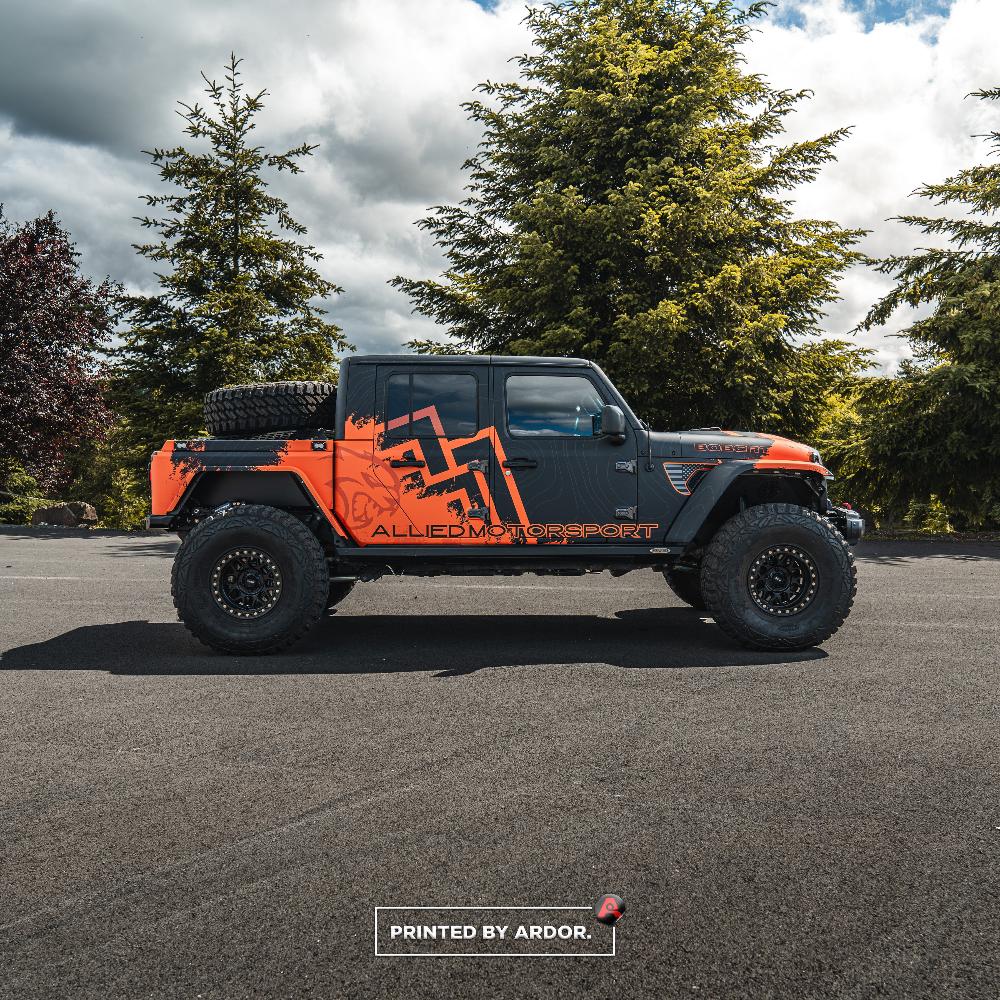 Side view of an Allied Motorsport Jeep Gladiator with a custom black and orange wrap, large off-road tires, and branding along the doors and bed.