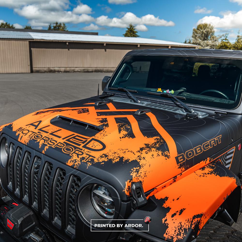 Hood view of an Allied Motorsport Jeep with orange and black custom wrap, featuring 'BOBCAT' branding and off-road styling, parked under blue skies.