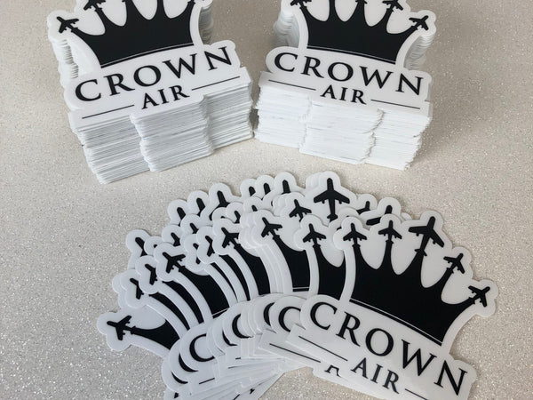 Stacks of white die-cut Crown Air logo stickers with a black crown and airplane design on a gray table