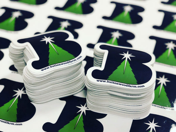 Two stacks of navy and green die-cut Buttonwood Farm logo stickers on a sheet of matching printed stickers