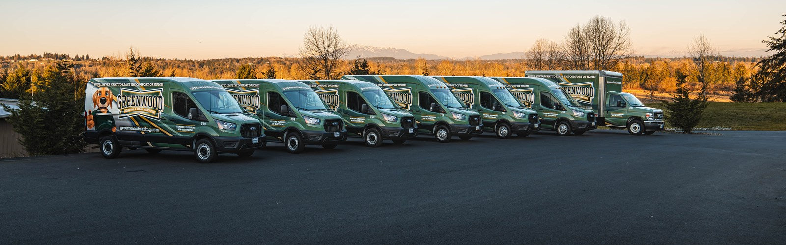 Fleet of green Ford Transit vans and a box truck in matching Greenwood Heating wraps, lined up at sunset