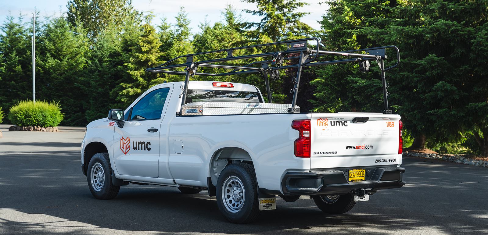 White Chevrolet Silverado work truck with UMC fleet lettering and logo graphics, upfitted with a black ladder rack